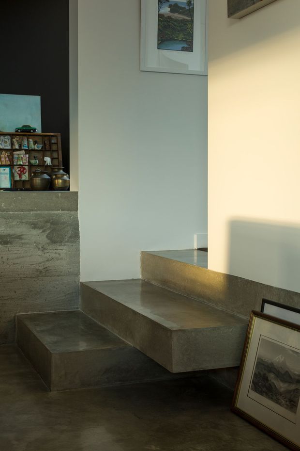 A moody interior view of a compact concrete stairway and raised platform. The space features raw grey concrete steps, landing and floor, pale plaster walls, warm directional sunlight, framed artwork leaning against the wall, and a small built-in display shelf with decorative objects. The composition emphasizes sculptural architectural elements and subtle material texture.