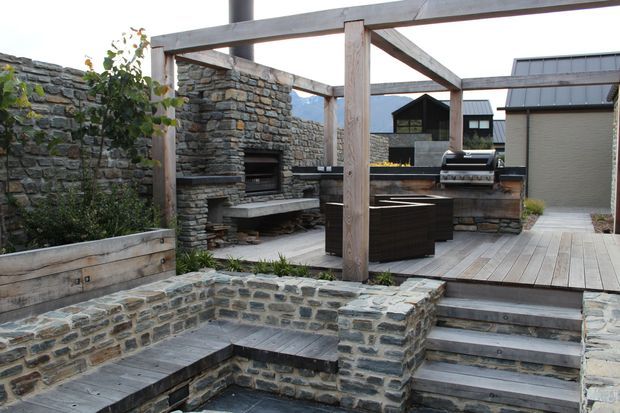 A landscaped outdoor entertaining area featuring a built-in stone fireplace, timber pergola structure, raised stone seating walls, timber decking, integrated bench seating, and an outdoor barbecue kitchen. The space has a rustic contemporary character and overlooks dark-clad outbuildings and mountainous scenery.