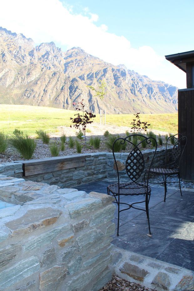 A landscaped outdoor terrace with dark stone paving, broad natural-stone steps, and a low stacked-stone retaining wall. Black metal lattice chairs sit beside the terrace, which overlooks a grassy open landscape and dramatic rugged mountain peaks. A dark-clad building edge is visible at right.