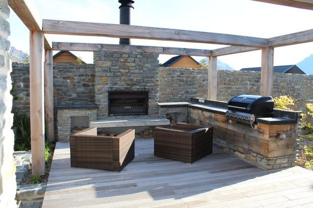 A sunlit outdoor entertaining area with a timber pergola over a paved deck. The space features a built-in stone fireplace, stone outdoor kitchen with barbecue, timber cabinetry, and two dark woven lounge chairs. Stone boundary walls and mountain scenery are visible beyond.