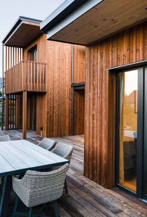 Exterior view of a contemporary multi-level building clad in warm vertical timber boards, with dark metal roof edges and window frames. A timber deck spans the foreground and includes an outdoor dining table with woven chairs. The composition highlights projecting balconies, covered outdoor areas, and large glazed openings.