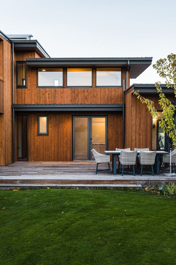 Exterior view of a contemporary two-storey timber-clad home with dark metal roof edges and window frames. A broad timber deck fronts the house and is furnished with an outdoor dining table and woven chairs, opening onto a lawn and garden.