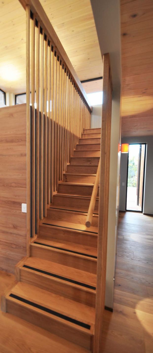 Vertical interior photograph focused on a timber staircase with open risers, solid wood treads, a matching handrail, and closely spaced vertical timber balusters. Timber wall and ceiling finishes continue through the stair hall, with a skylight and glazed doorway providing natural light. A hallway or living area is visible beyond the stair.
