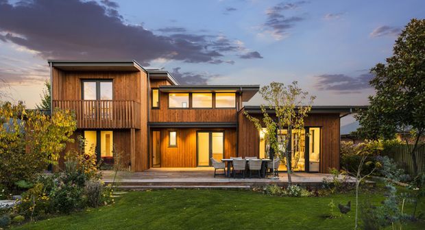 A contemporary multi-level timber-clad home viewed from a landscaped garden at dusk. The house features several offset flat-roofed volumes, large glazed openings, a projecting upper balcony, and a timber deck with outdoor dining furniture. Warm interior lighting contrasts with the cool evening sky.