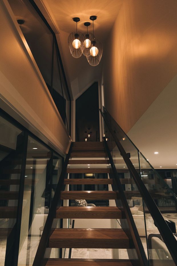 Interior view looking up a contemporary open-riser staircase with thick dark timber treads, black metal stringers, and clear glass balustrades. The stair rises through a tall double-height void, with black-framed glazing, warm timber wall and ceiling finishes, and sculptural wire pendant lights. A living area is visible beneath and beyond the staircase.