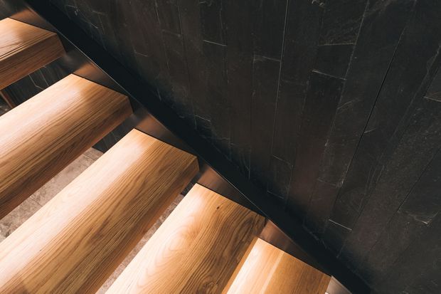 Close-up view of a contemporary stairway with thick floating timber treads, warm integrated lighting beneath the steps, and a dark vertically panelled wall alongside. The composition highlights the contrast between the natural wood grain and the near-black wall finish.