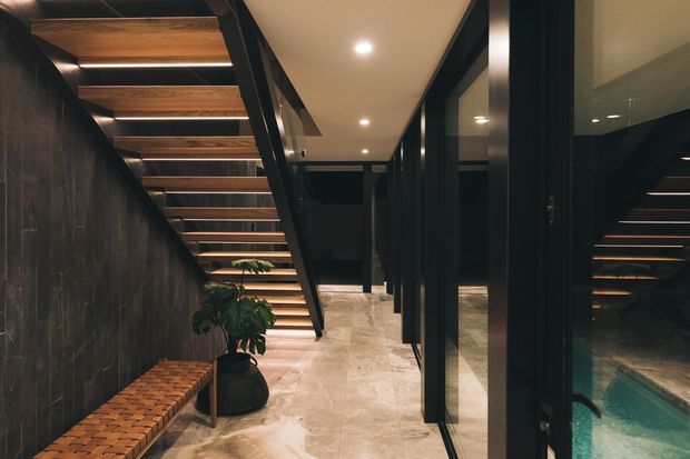 A dramatic contemporary interior corridor beneath an open-riser staircase. Dark timber wall panelling and black steel framing contrast with warm timber stair treads, pale stone flooring, recessed ceiling lights, and full-height glazed partitions. A built-in-looking timber bench and potted plant sit along the left wall, while a swimming pool is visible through the glazing on the right.