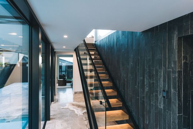 Modern interior hallway with a floating timber staircase, slim black handrail, and clear glass balustrade. The stair is set against a dark vertically veined stone feature wall, with extensive floor-to-ceiling glazing along the corridor and a skylight or roof opening above.