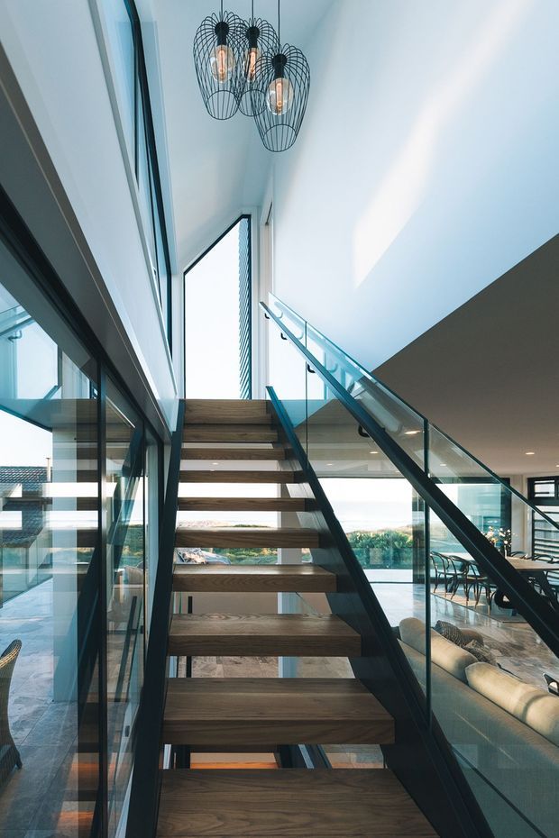 A bright contemporary interior view looking up an open-riser staircase with dark timber treads, slim dark metal stringers, and clear glass balustrades. The stair connects a double-height living and dining area, with expansive glazing, a pale ceiling, and three black wire-cage pendant lights. A sofa and dining setting are visible beside the stair, while the glazing reveals a distant outdoor landscape.