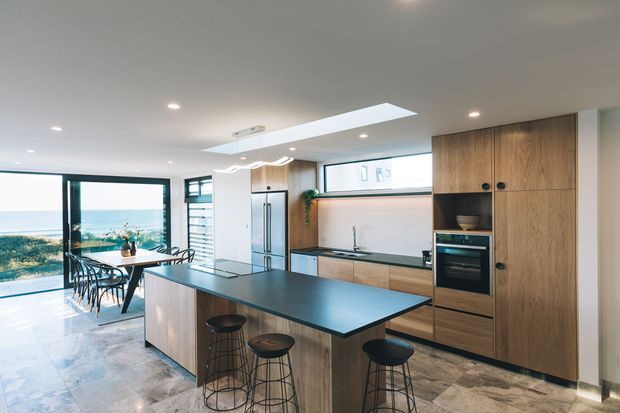 Bright open-plan coastal kitchen and dining area with a large dark benchtop island, timber cabinetry, integrated stainless-steel appliances, skylight, and expansive glazing overlooking the sea.