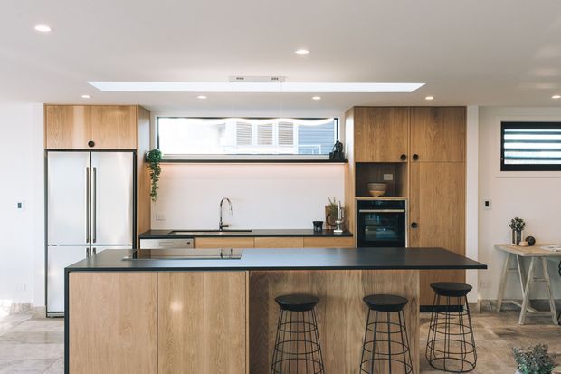 A bright contemporary kitchen featuring a large timber island with dark stone benchtop and seating for three, full-height timber cabinetry, integrated appliances, a stainless-steel refrigerator, open shelving, and a wide skylight. A small dining area is visible to the right.