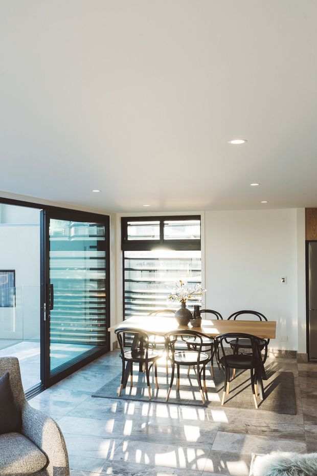 Bright contemporary dining area with a large timber dining table and dark bentwood-style chairs, positioned beside full-height glazed sliding doors and horizontal louvre windows. Natural sunlight creates strong linear shadows across a pale stone-tiled floor, with a neutral white interior and glimpses of a kitchen at right.