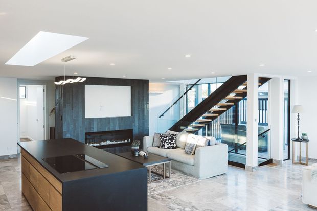 Bright open-plan contemporary interior combining a kitchen and living room. A large dark island with integrated cooktop dominates the foreground, while a pale sectional sofa faces a dark timber feature wall with a fireplace and television recess. A sculptural open-riser staircase with dark timber stringers and glass balustrading rises beside floor-to-ceiling glazing. A skylight, recessed lighting, marble-look flooring, and clean white walls create an airy, refined space.