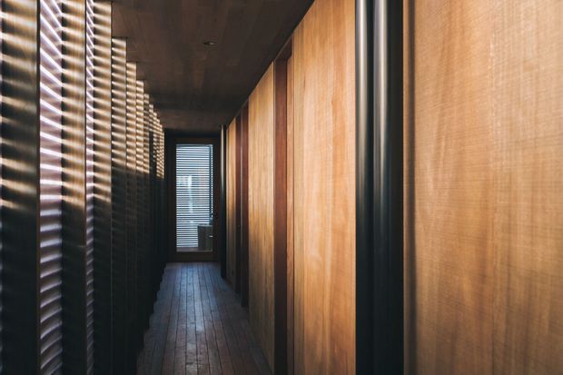 A narrow, dark architectural corridor lined with warm timber wall panels and dark vertical metal or timber slats. The corridor has a timber floor, a dark ceiling, and a small louvred window at the far end, creating a dramatic interplay of light and shadow.