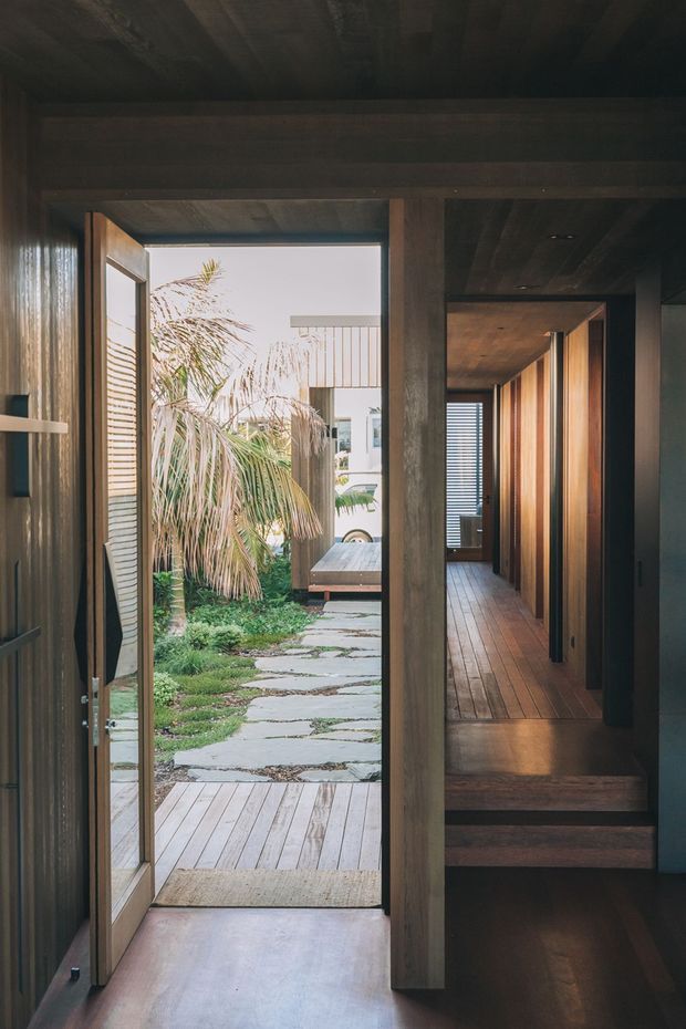 A dark, timber-lined interior entryway opens through a glazed timber door to a landscaped courtyard. The space features exposed timber ceiling and wall surfaces, warm wood flooring, a short flight of timber steps, and a long corridor with vertical timber doors and panels. Natural daylight and greenery beyond the doorway contrast with the moody interior.