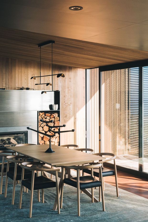 Warm, refined dining room with a large timber dining table and matching wooden dining chairs arranged on a rug. The room features extensive vertical timber wall and ceiling lining, a sculptural black pendant light, a built-in fireplace with stacked firewood, and expansive glazing with vertical blinds. Natural sunlight creates strong linear shadows across the timber surfaces and floor.