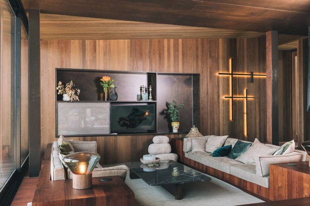 A sophisticated, wood-lined living room with floor-to-ceiling glazing along one side, dark timber ceiling and walls, integrated media and display cabinetry, a large sectional sofa, glass coffee table, and a sculptural illuminated wall feature. The room has a warm, moody, refined character with contemporary furnishings and layered timber finishes.