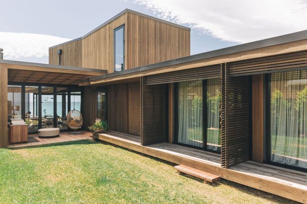 Modern single-storey courtyard-style residence with warm vertical timber cladding, expansive glazed sliding doors, deep timber verandah decking, and horizontal timber privacy screening. A taller box-like upper volume rises behind the low wing, while an outdoor seating area connects to the lawn and garden courtyard.