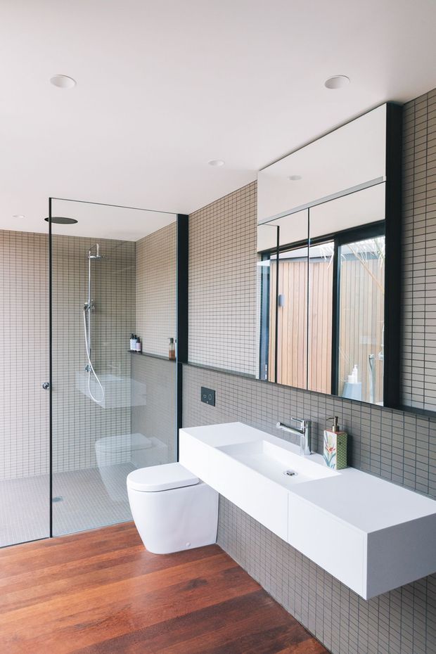Contemporary bathroom with a walk-in glass shower, wall-mounted white toilet, floating double vanity, large mirror, and small-format grey tiled walls. The room combines warm timber flooring with crisp white fixtures and dark-framed glazing, creating a clean, bright and refined feel.