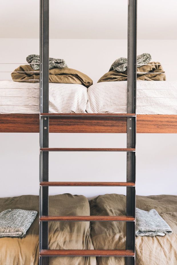A close-up, symmetrical view of a bunk bed with a central ladder. The bed has dark metal vertical rails and warm reddish-brown timber rungs and framing, with neutral beige and grey bedding against a plain white wall. The composition highlights the built-in ladder and the two sleeping levels.