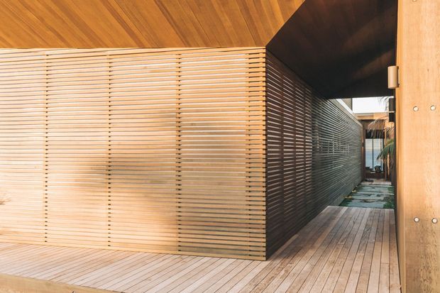 A narrow, contemporary timber-clad passage or courtyard walkway with finely spaced horizontal timber screening forming a private enclosure. The warm timber ceiling, slatted walls, timber decking, and dark overhead section create a strong linear perspective toward a partially visible exterior courtyard.