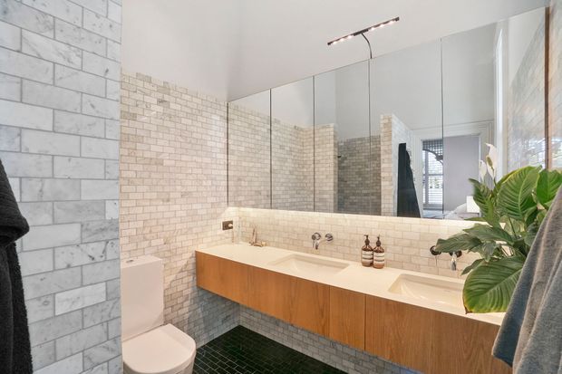 Bright contemporary bathroom featuring a long floating timber vanity with integrated white basins, a large mirrored wall, pale brick-pattern wall tiles, and dark green floor tiles. A toilet and leafy plant are visible, with natural light entering from the right.