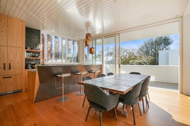Bright open-plan kitchen and dining area with warm timber cabinetry and flooring, a dark stone kitchen island with bar seating, a large timber dining table, upholstered dining chairs, pendant lights, and expansive windows opening toward an outdoor terrace. The space has a refined mid-century-inspired character with abundant natural light.