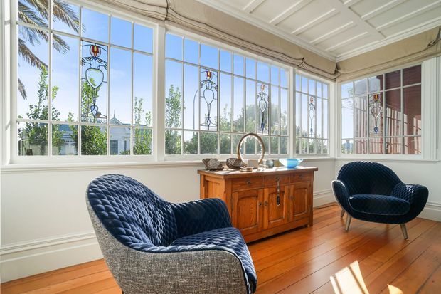 Bright enclosed sunroom with expansive multi-pane windows, decorative stained-glass inserts, white wall panelling and coffered ceiling, warm timber flooring, a traditional wooden sideboard, and two sculptural navy upholstered lounge chairs. Palm foliage and a garden are visible outside.