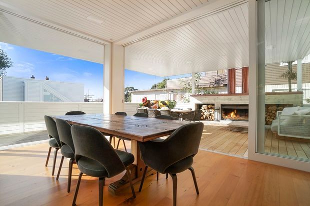 Bright, open-plan dining and outdoor entertaining area with a large timber dining table and upholstered chairs in the foreground. The space opens onto a covered timber deck with a built-in fireplace, stacked firewood, outdoor seating and glazing reflecting the interior. White-lined ceilings, pale walls, extensive openings and warm timber flooring create an airy coastal feel.