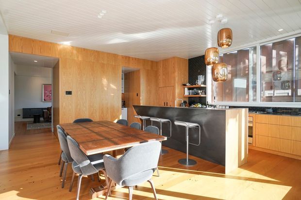 A bright open-plan kitchen and dining area with extensive warm timber wall panelling and cabinetry, a dark stone kitchen island with integrated bar seating, and a large timber dining table surrounded by upholstered dining chairs. Copper pendant lights hang above the island, while expansive windows bring in strong natural light. A glimpse of an adjacent living area is visible through the opening at left.