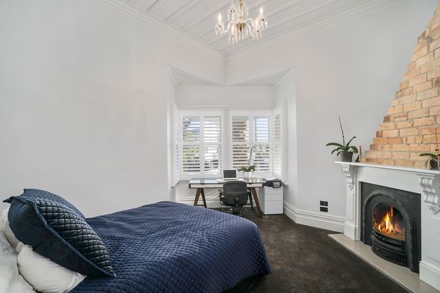 Bright, refined bedroom with a navy quilted bed and matching pillows, a compact desk and office chair positioned in a bay window, white plantation shutters, ornate ceiling detailing, a crystal chandelier, and a traditional exposed-brick fireplace with a white mantel. The room has dark carpeting and a calm, classic character.