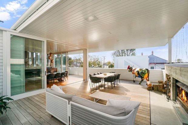 Bright, expansive covered outdoor living and dining terrace adjoining a glazed interior. The space features timber decking, a large white slatted ceiling, floor-to-ceiling sliding glass doors, a dining table with chairs, outdoor lounge seating, and a built-in masonry fireplace with a fire. The terrace overlooks neighbouring residential buildings and trees, creating an airy indoor-outdoor entertaining area.