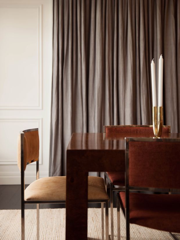 A refined dining area with a dark polished wood dining table and coordinating upholstered dining chairs. Floor-to-ceiling taupe-brown curtains frame a white decorative wall with panel moulding, while a pair of white taper candles in a gold-toned holder provides a restrained accent. A pale textured rug softens the dark timber floor, creating an elegant, warm and intimate composition.