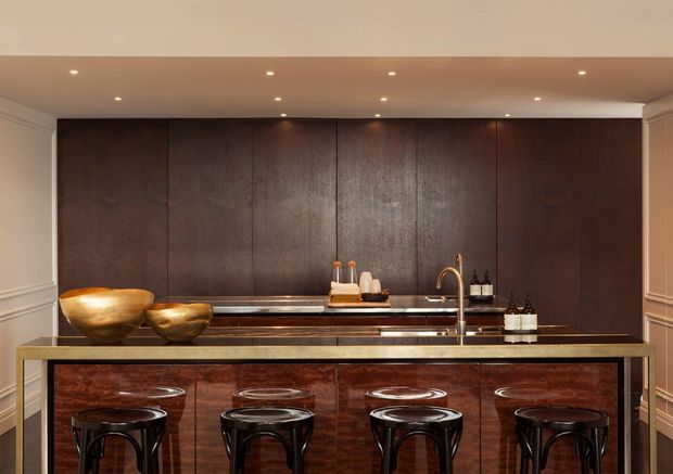A sophisticated built-in home bar with dark wood wall panelling and a matching richly figured wood island. The bar features a long stone or dark composite benchtop, integrated sink with brass-toned tapware, open shelving or recessed display area, four black bar stools, and warm decorative lighting. Classic wall mouldings frame the space, creating a refined, luxe atmosphere.