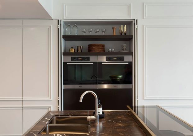 Close-up view of a refined kitchen featuring white raised-panel cabinetry, a dark marble or stone island benchtop with integrated double sink and curved chrome tap, black built-in double ovens, open shelving with glassware and crockery, and a flush-mounted cooktop. The space has a classic, tailored fit-out with strong dark-and-light contrast.