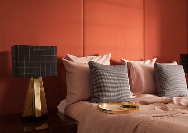 A warmly lit bedroom with a richly terracotta-red upholstered or paneled feature wall, layered blush and dusty-pink bedding, and contrasting black-and-white check cushions. A sculptural brass-gold table lamp with a dark grid-patterned shade sits on a dark bedside table, while a round gold tray rests on the bed. The styling is intimate, glamorous, and high-contrast with strong jewel-box colour.