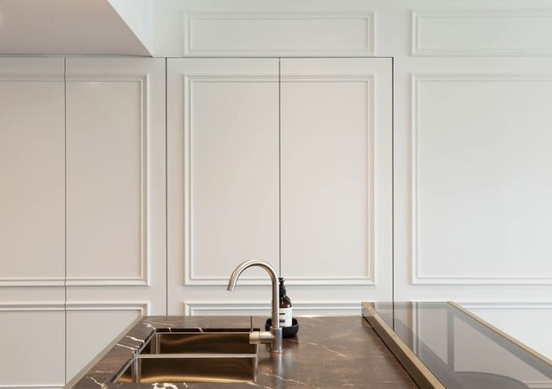 Close-up view of a refined kitchen with white raised-panel cabinetry and matching decorative wall panelling. A dark brown marble or natural stone benchtop surrounds a stainless-steel undermount sink and chrome tap, with a glass splashback or glazed divider and a black glass cooktop visible at right. The composition is bright, symmetrical, and detail-focused.