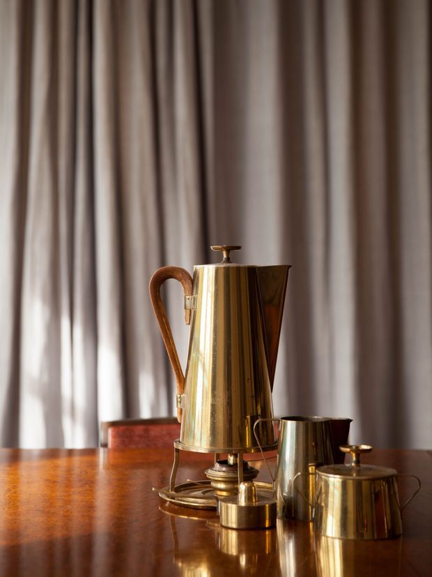 A close-up still-life of a polished brass or copper coffee and tea service arranged on a glossy dark wood table. The set includes a tall lidded urn with a curved wooden handle, a smaller metal pitcher, and additional lidded vessels. Floor-to-ceiling pleated curtains form a softly lit neutral backdrop, creating a warm, refined vintage atmosphere.
