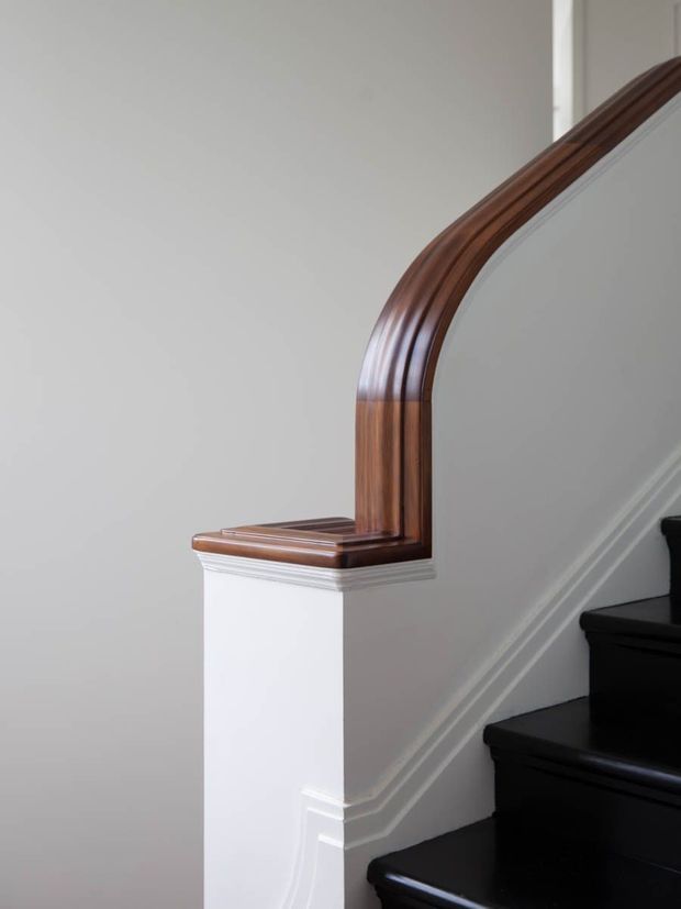 Close-up architectural detail of a traditional staircase, featuring glossy black stair treads, white painted stair walls and trim, and a richly stained curved timber handrail with decorative moulded detailing.