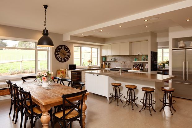 Open-plan kitchen and dining area with a large rustic timber dining table, dark cross-back chairs, a central white island with a stone-look benchtop, and four industrial-style stools. Cream cabinetry, stainless-steel appliances, pendant and recessed lighting, and broad windows create a bright, warm family space.