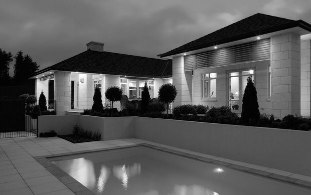 A black-and-white dusk exterior photograph of a refined single-storey residence surrounding a rectangular swimming pool. The home features pale masonry walls, dark tiled hipped roofs, large glazed doors and windows, horizontal louvred detailing, recessed exterior lighting, and formal clipped planting in raised rendered planters.