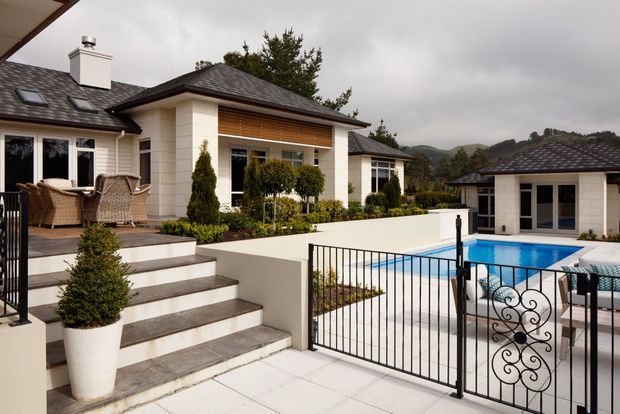 A refined single-storey residential exterior with a dark tiled hipped roof, cream masonry walls, timber-look horizontal cladding accents, broad outdoor steps and terrace, and a gated swimming pool courtyard. A covered outdoor dining area and landscaped planting sit beside the main house, with wooded hills visible in the distance.
