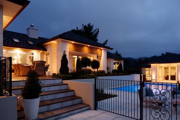 A luxury contemporary residence at dusk, viewed from an elevated outdoor terrace or courtyard. The home features pale rendered walls, dark tiled pitched roofs, warm timber-look soffit and wall cladding, extensive glazing, landscaped planting, broad steps, decorative metal fencing, and a brightly lit swimming pool courtyard.