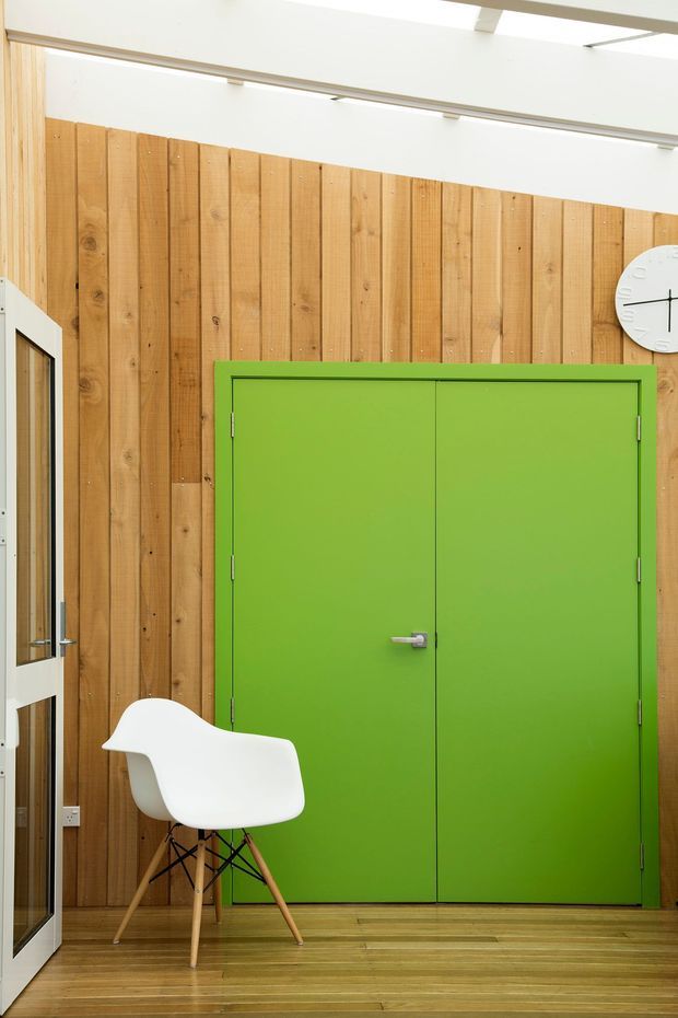 Bright contemporary interior featuring a full-height vertical timber wall, a vivid green double door, pale timber flooring, white ceiling beams, and a sculptural white chair near a glazed white-framed door. The image is focused on a bold internal entry or circulation space with clean modern detailing and strong colour contrast.