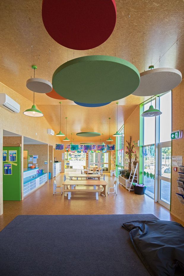 Bright, colourful communal interior resembling a childcare, school, or community learning space. The room has a warm wood-fibre or timber-toned wall and ceiling finish, polished warm-toned flooring, large windows and glazed entry doors, rows of communal tables and chairs, vivid green built-in cabinetry, colourful artwork and displays, and multiple suspended circular acoustic ceiling panels in green, red, blue, and grey. A dark carpeted foreground seating or activity area adds contrast.