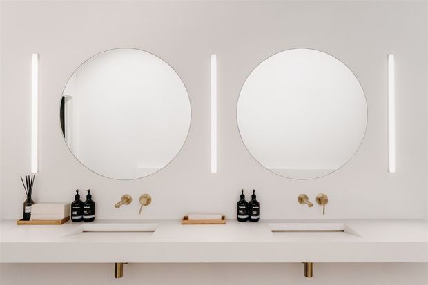 Bright, minimalist double vanity bathroom with two large round frameless mirrors, slim vertical wall lights, white integrated basins and benchtop, and simple black and brass fixtures and accessories. The composition is symmetrical and clean, with a pale neutral palette and concealed or minimal storage.