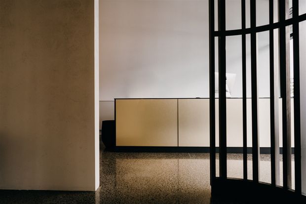 A minimalist interior with a warm beige plaster wall, pale built-in cabinetry, dark trim, and a black-framed glazed partition creating a strong geometric screen. The polished speckled flooring reflects the cabinetry and partition, while the restrained palette and clean lines create a refined, contemporary atmosphere.