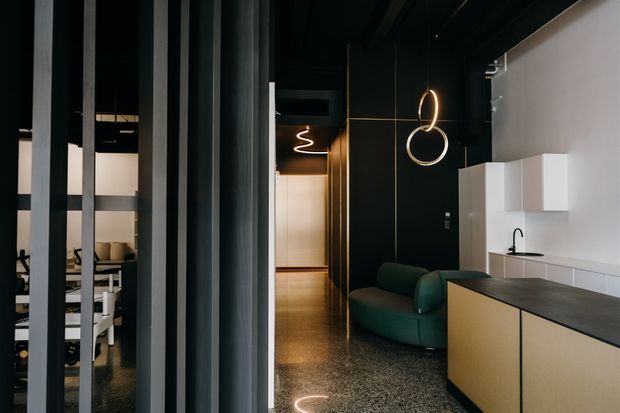 A dark, refined contemporary interior viewed through black vertical partitions. The foreground shows a compact kitchen with pale integrated cabinetry, white wall cupboards, a dark benchtop and black tap. Beyond it is a deep green curved lounge chair, a polished speckled floor, and sculptural pendant lighting. A partially screened dining or workspace area is visible to the left.
