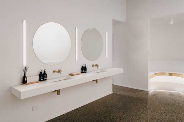A bright, refined bathroom with a long floating double vanity, two round mirrors, vertical illuminated wall lights, integrated basins, and brass tapware. The neutral white fit-out contrasts with a speckled terrazzo-like floor, while an arched opening leads to a partially visible curved freestanding bath.