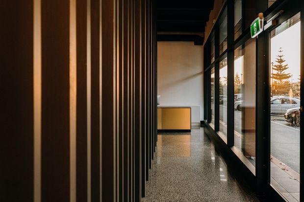 A narrow, dark-toned commercial or institutional corridor with a dramatic vertical timber-slat wall on the left, black-framed full-height glazing on the right, and a polished terrazzo-like floor. Warm light illuminates a simple reception or display counter at the far end, while the glazing reveals a street-facing exterior.